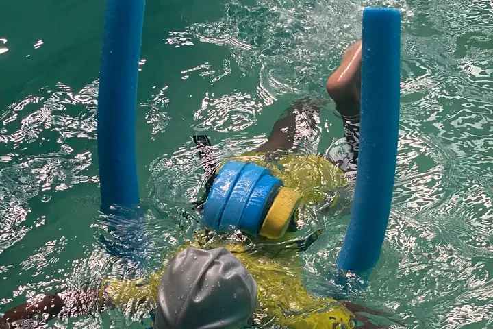 A young swimmer in a cap, seen from above, floating between two pool noodles
