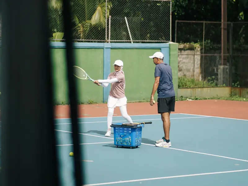A coach standing beside a player as she hits a backhand on a hard court