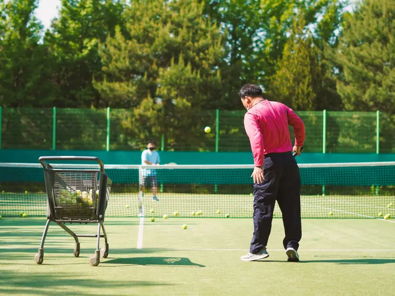 A coach with a ball cart feeding balls to a player across the net