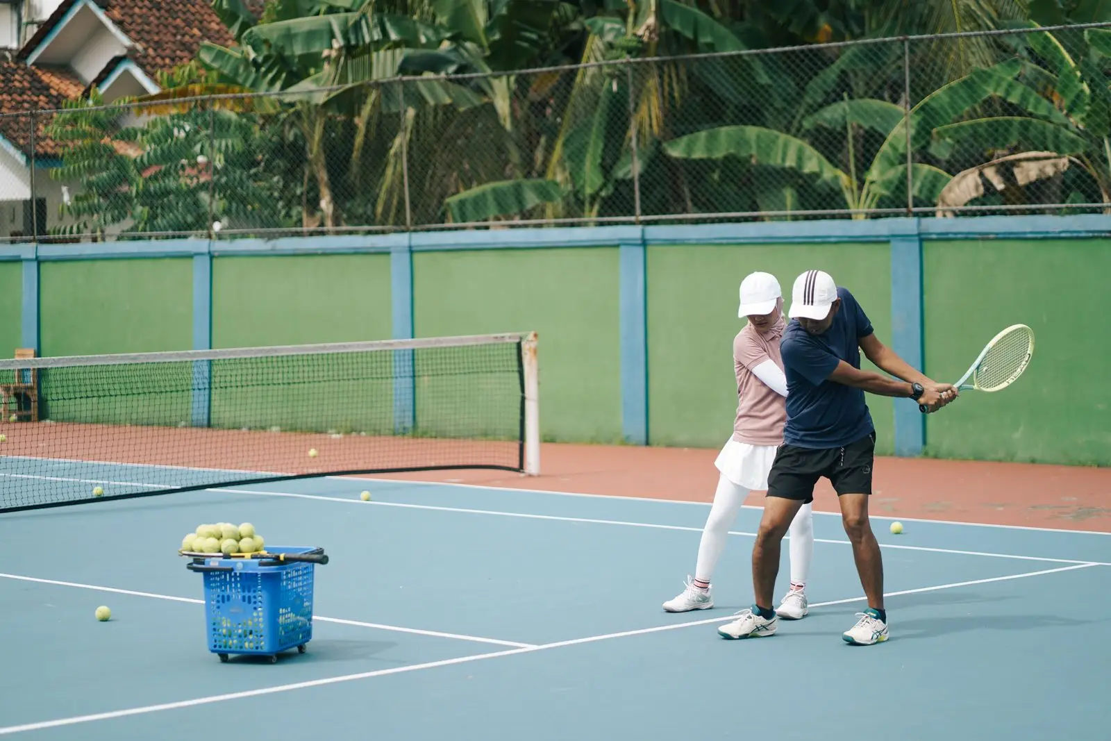 A tennis coach and an adult player working on a two-handed backhand together on an outdoor hard court